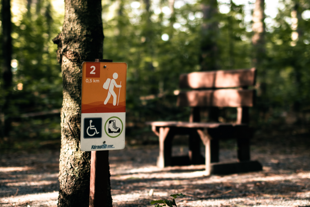 En arrière plan, une forêt avec un sentier de marche. Au premier plan, une affiche indiquant la position dans le sentier et le pictogramme de l'accessibilité.