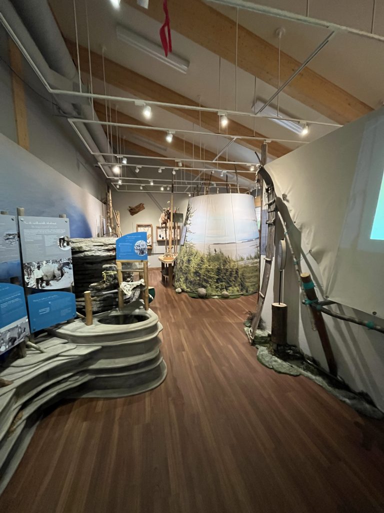 Exposition avec des objets et installations culturelles innues, dans une salle avec un éclairage tamisé et un plafond en bois.