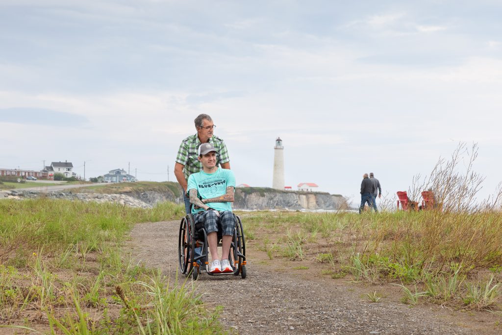 Un homme pousse un autre homme en fauteuil roulant sur un chemin de terre près d’un phare en bord de mer, sous un ciel nuageux. Des bâtiments et des chaises rouges se trouvent à l'arrière-plan.