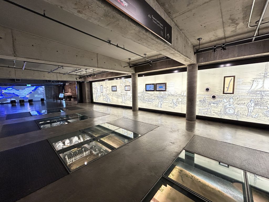 Salle moderne et interactive de Pointe-à-Callière avec une exposition historique illustrée sur les murs, des vitrines au sol et des écrans numériques.