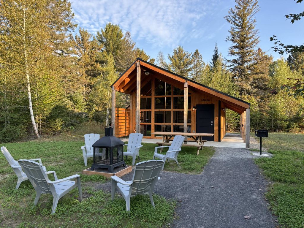 Mini-chalet en bois avec de grandes fenêtres et un foyer extérieur, entouré de chaises Adirondack. Sentier accessible menant à l'entrée, situé au cœur de la nature aux Chalets Lanaudière.