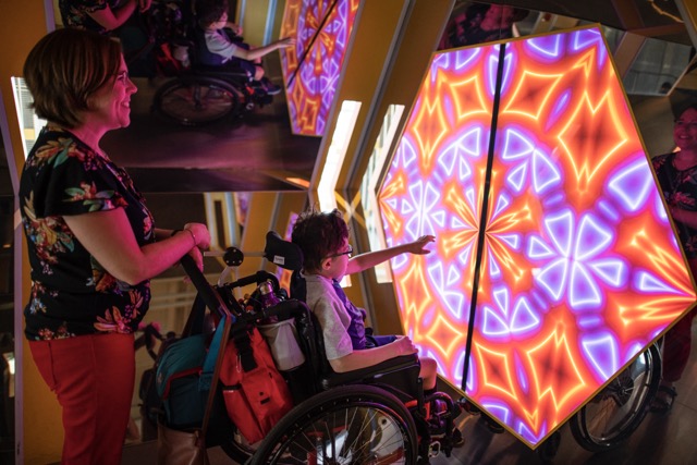 Enfant en fauteuil roulant pointant une structure interactive lumineuse, accompagné d’un adulte, dans une activité accessible au Québec.